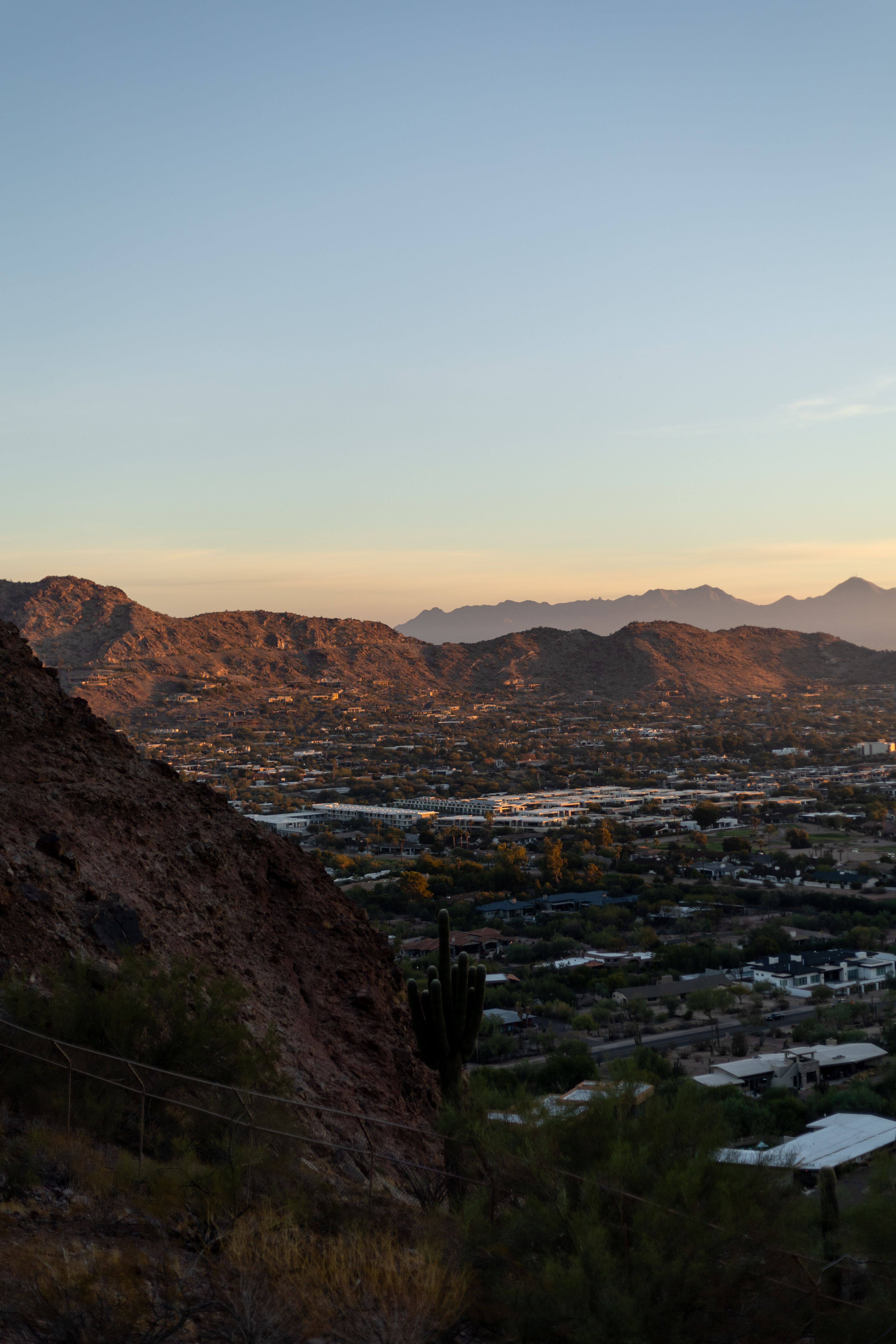 Warm evening light across desert mountains and a Southwest city, viewed from a rocky hillside.